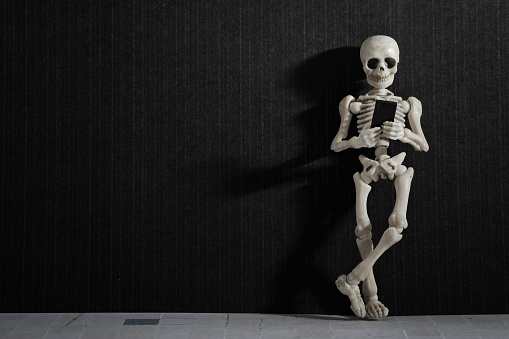 A skeleton leaning on a black wall with smartphone in his hand