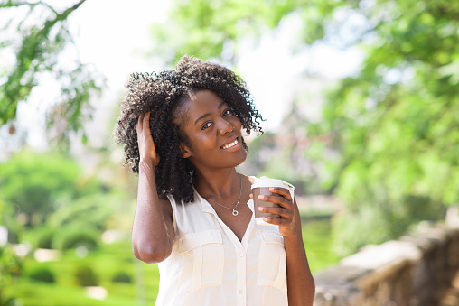 Portrait of happy young African-American woman wearing blouse drinking coffee, tousling hair and smiling outdoors. Drink and break concept