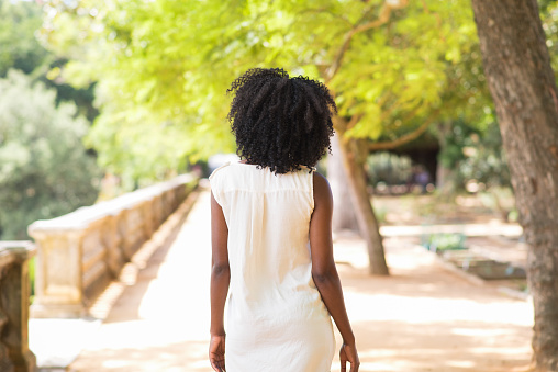 Rear view of young African-American woman wearing white dress walking in park. Leisure and recreation concept