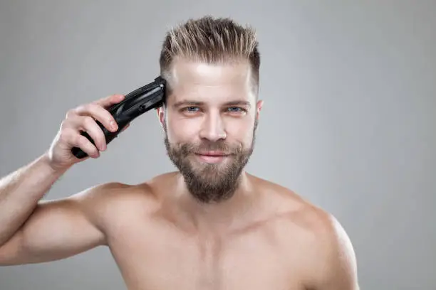 Handsome bearded man cutting his own hair with a clipper