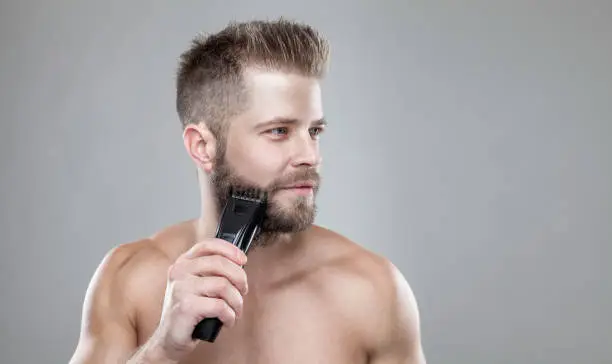 Handsome young bearded man trimming his beard with a trimmer