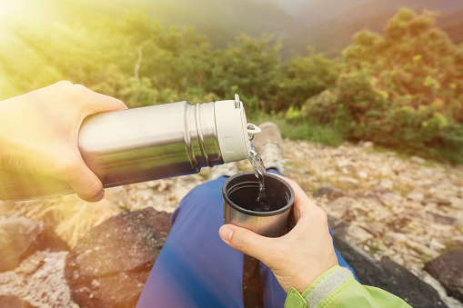 Steel thermos in hand. Pour a hot drink into a mug. A glass in his hand with water. Bask in an herbal drink from a thermo bottle. Winter hike in the woods. High quality photo
