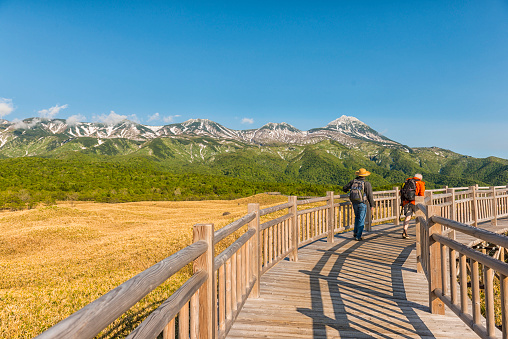 Scenic view on long boardwalk over wetland and two men walking, Shiretoko Five lakes. In backgrounds blue sky and snowcapped mountains, Hokkaido, Japan
