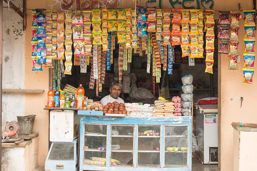 Kambeswarji: Indian vendor sits in shop with chips & namkeen hanging in background and other products. pakazing namkeen and chips are big sale in india
