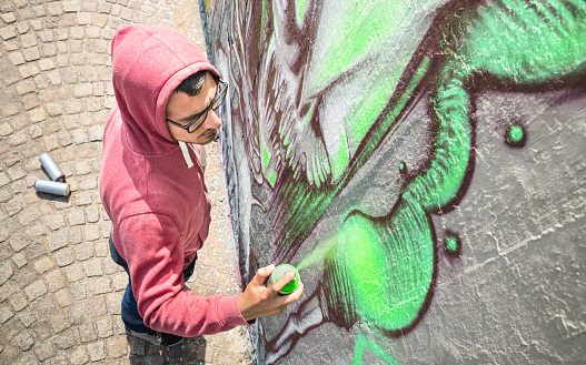 Street artist painting colorful graffiti on generic wall - Modern art concept with urban guy performing and preparing live murales with green aerosol color spray - Sunny afternoon neutral filter