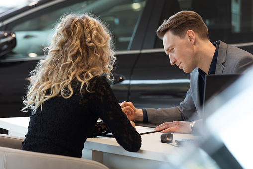 Car salesperson talking with female customer while sitting at desk in showroom