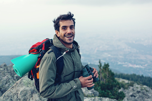 Young smiling man standing on the mountain top, holding binoculars and smiling at camera