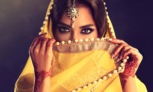 Close up portrait of young indian woman is covering part of the face by yellow cloth. Bright make up, long eyelashes and tender look. Henna tattoos(mehndi) and gilded manicure on hands.