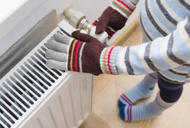 A child in woolen gloves and a sweater warms his hands near the heater.