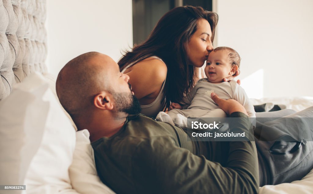 Parents with their newborn baby boy on bed Parents with their newborn baby boy on bed at home. Woman kissing her son sitting with father. Baby - Human Age Stock Photo