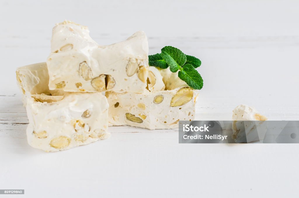 Delicious Italian festive torrone or nougat Delicious traditional Italian festive torrone or nougat with nuts on white wooden background with place for text. Soft nougat blocks with almonds with fresh mint leaves. Italian sweets. Copy space. Affectionate Stock Photo