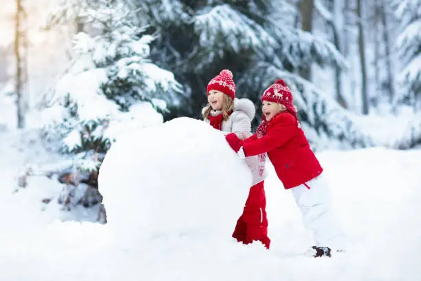 Children build snowman. Kids building snow man playing outdoors on sunny snowy winter day. Outdoor family fun on Christmas vacation. Boy and girl play snow balls. Winter clothing for baby and toddler.