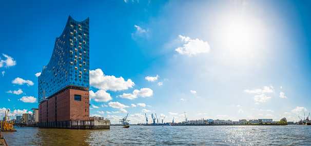 Bright sunburst in blue skies over the cranes and docks of the Port of Hamburg and the iconic sail of the Elbphilharmonie on the shores of HafenCity, the redeveloped waterfront of Germany’s vibrant second city.