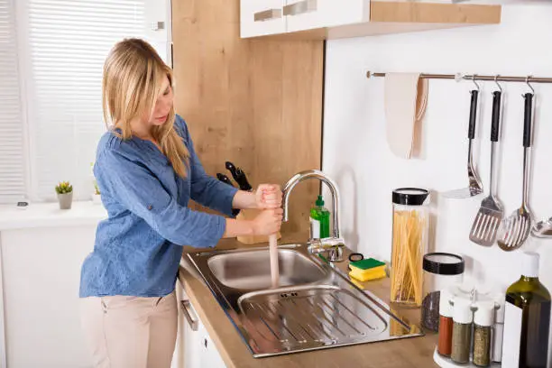 Young Woman Using Plunger In Blocked Kitchen Sink To Unclog Drain At Home