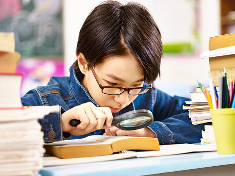 asian pupil with glasses using a magnifier to enlarge the words in a thick book.