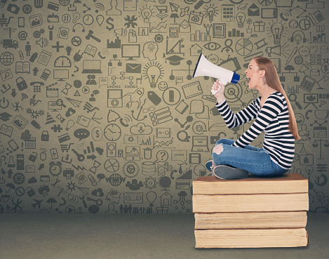 Young female student sitting on stack of books and speaking in megaphone