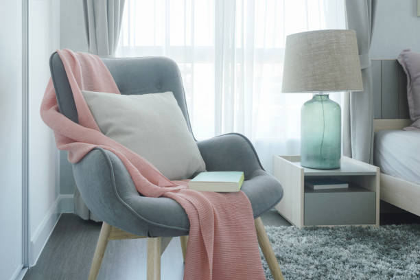 Gray easy armchair with pink scarf, pillow and book next to bed in the bedroom stock photo