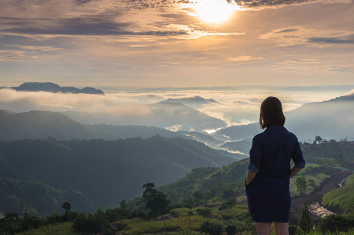 Woman standing with hand in her pockets while looking at the panoramic view of mountain range and mist under the sunset