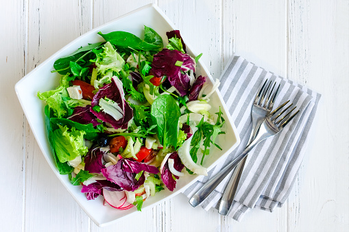 Fresh summer green salad mix with salad lettuce, spinach, fennel, celery, tomatoes, radish, olives, chicory and arugula in white bowl with forks on wooden background. Healthy eating concept. Top view.