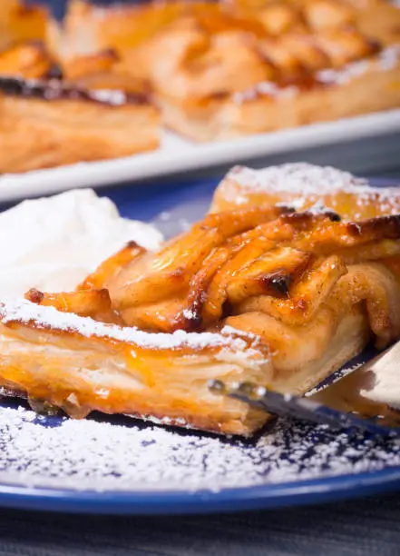 Piece of an apple tart baked in puff pastry with an apricot glaze dusted in powdered sugar and served with fresh whipped cream.