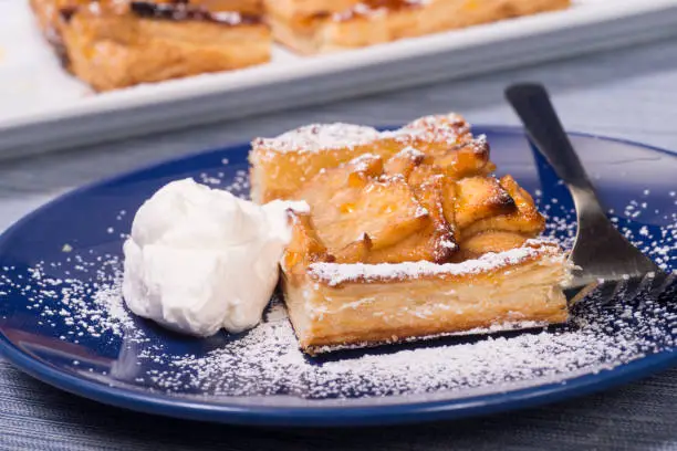 Piece of an apple tart baked in puff pastry with an apricot glaze dusted in powdered sugar and served with fresh whipped cream.