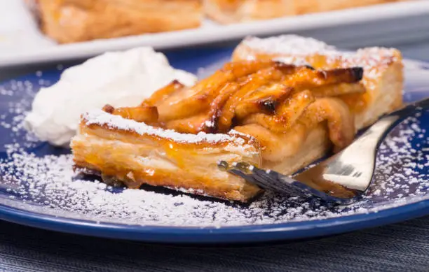 Piece of an apple tart baked in puff pastry with an apricot glaze dusted in powdered sugar and served with fresh whipped cream.