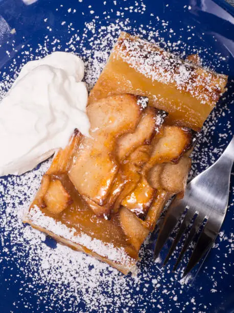 Piece of an apple tart baked in puff pastry with an apricot glaze dusted in powdered sugar and served with fresh whipped cream.