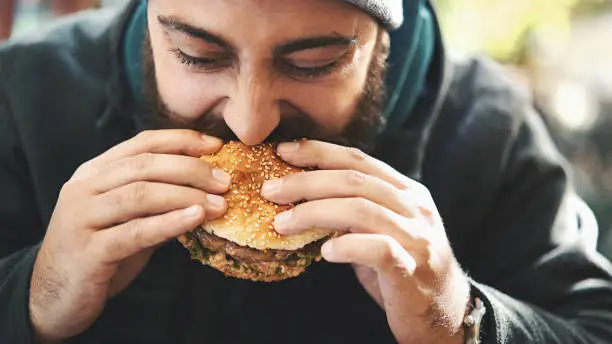 Closeup front view of a mid 20's man biting on a juicy burger outdoors. He has brown beard and mustache. He's wearing winter jacket and a cap.