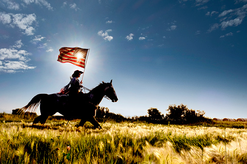 Young woman riding horse carrying American flag in grassy green field with mountains in the background.