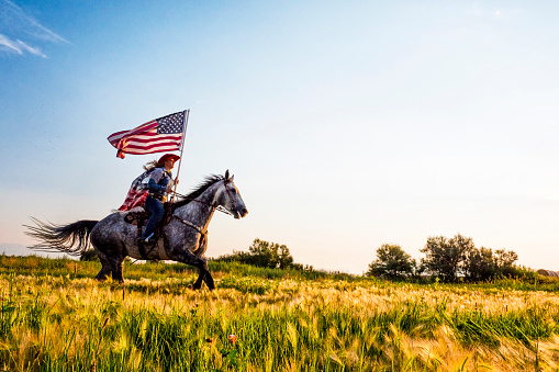 Young woman riding horse carrying American flag in grassy green field with mountains in the background.