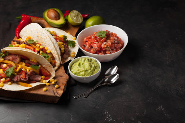 istock Three Mexican tacos with marbled beef, black Angus and vegetables on wooden Board on a dark stone background. Mexican dish with sauces guacamole and salsa in bowls. Top view with copy space 859504046