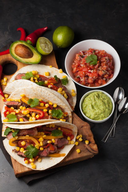 istock Three Mexican tacos with marbled beef, black Angus and vegetables on wooden Board on a dark stone background. Mexican dish with sauces guacamole and salsa in bowls. Top view 859504110