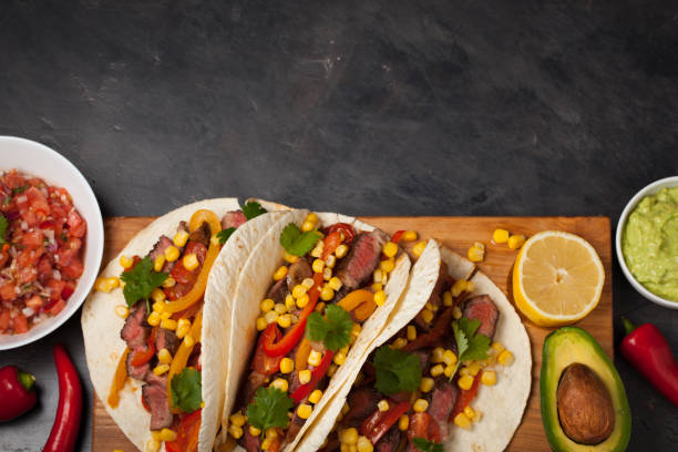 istock Three Mexican tacos with marbled beef, black Angus and vegetables on wooden Board on a dark stone background. Mexican dish with sauces guacamole and salsa in bowls. Top view with copy space 859504176