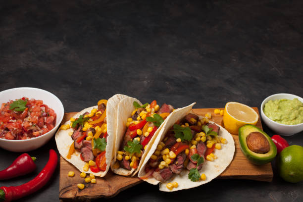 istock Three Mexican tacos with marbled beef, black Angus and vegetables on wooden Board on a dark stone background. Mexican dish with sauces guacamole and salsa in bowls. Top view with copy space 859504194