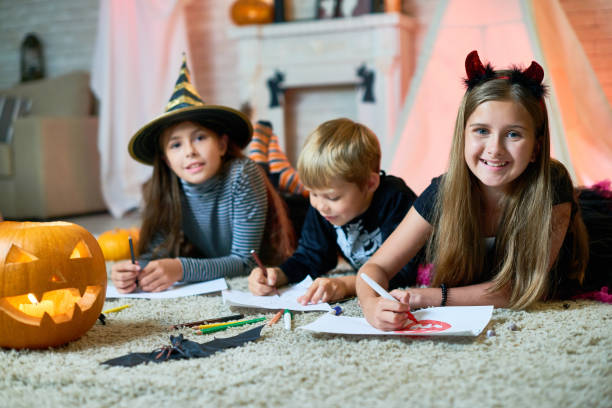 Creative Children at Halloween Party Group portrait of cheerful children wearing Halloween costumes gathered together at cozy living room decorated for Halloween and drawing colorful pictures, girls looking at camera with wide smiles coloring photos stock pictures, royalty-free photos & images
