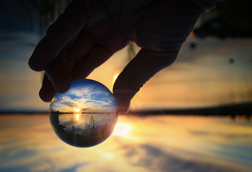 Fingers holding transparent glass ball reflecting the sunset on the water of Cold Lake, Alberta
