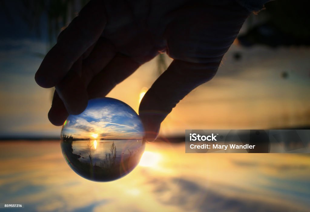 Transparent glass ball reflecting a sunset on the lake Fingers holding transparent glass ball reflecting the sunset on the water of Cold Lake, Alberta Looking Through Window Stock Photo