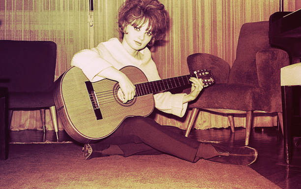 Vintage black and white toned photo from the sixties of a young woman playing guitar sitting in the living room