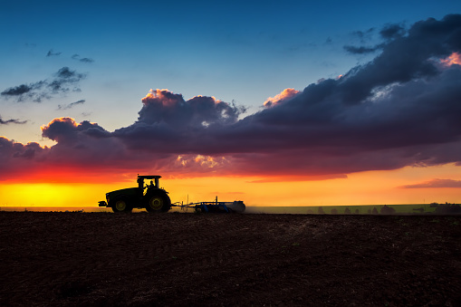 Farmer in tractor preparing land with seedbed cultivator, sunset shot. Silhouettes.