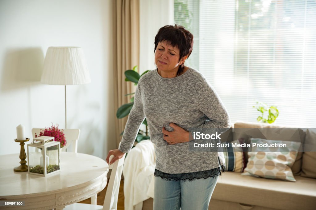 Mature woman suffering from stomach ache at home Mature woman suffering from stomach ache at home. Grabbing and squeezing belly with hand, feeling exhausted, standing in living room. Expressing pain on face Abdomen Stock Photo