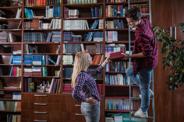 Smiling teenage boy standing on the ladder in a library and giving a book to his female friend.
