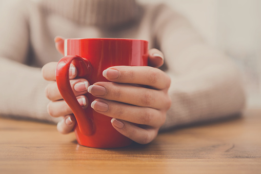 Woman holding cup of tea while sitting at wooden table in cozy coffee shop, shallow depth of field. Girl enjoying mug with hot drink at urban cafe and warming hands