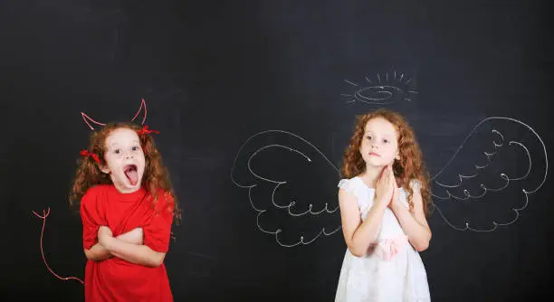 Two kids characters. Angry girl with imp horns and tail and a angel girl wings drawn on a blackboard.
