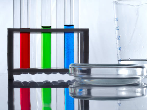 provette con liquido blu, verde e rosso in un portatubi, una tazza di misura e una piastra di petri su un tavolo bianco e sfondo bianco. la scrivania del biologo. - test tube biologist laboratory beaker foto e immagini stock