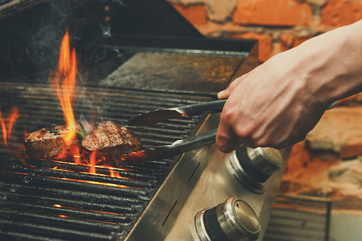 Man cooking meat steaks on professional grill outdoors. Male hand with tongs flipping beefsteaks on open fire