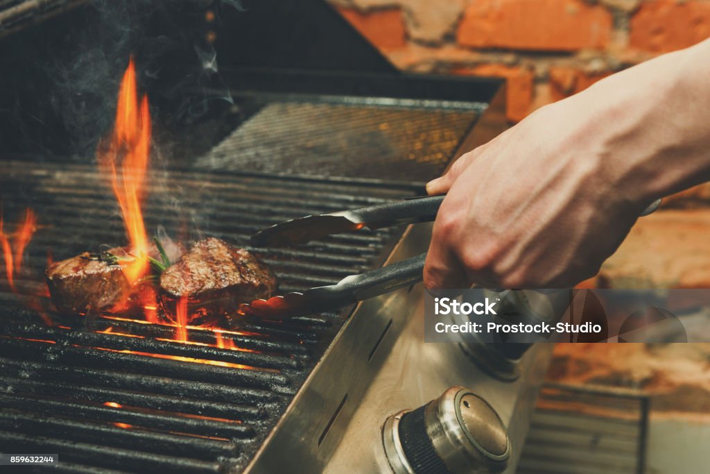 Man cooking meat steaks on professional grill outdoors Man cooking meat steaks on professional grill outdoors. Male hand with tongs flipping beefsteaks on open fire Barbecue Grill Stock Photo