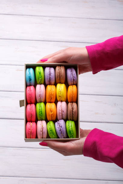 istock Hands holding gift box with delicious macaroons on white wooden background, top view. 859642440