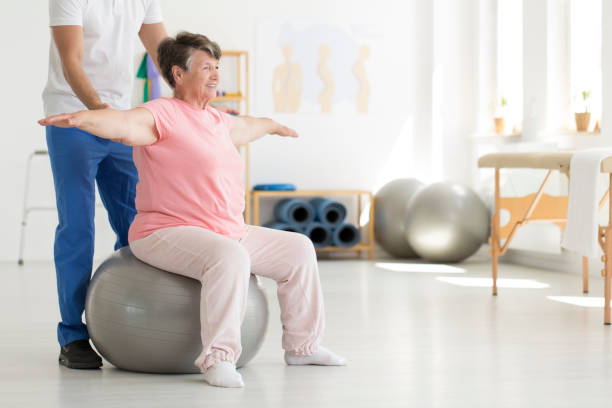 Ward sitting on fit ball Elderly senior ward trying to maintain balance while sitting on a grey fit ball while being supported by physiatrist hip replacement stock pictures, royalty-free photos & images