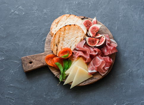 Delicious appetizers for wine or a snack - prosciutto, figs, bread, cheese on a rustic wooden cutting board. On a gray background, top view