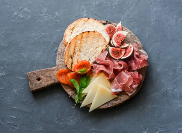 Delicious appetizers for wine or a snack - prosciutto, figs, bread, cheese on a rustic wooden cutting board. On a gray background, top view